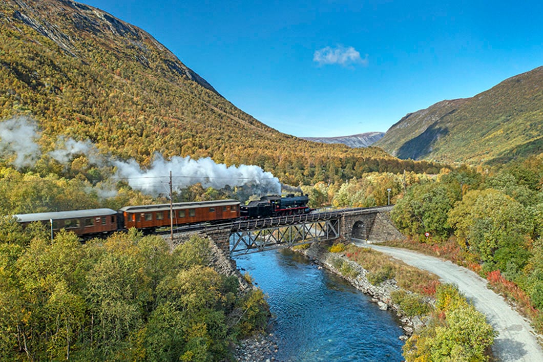 Et gammelt damptog på vei over en bru i et vakkert terreng med elv, åssider og skog.