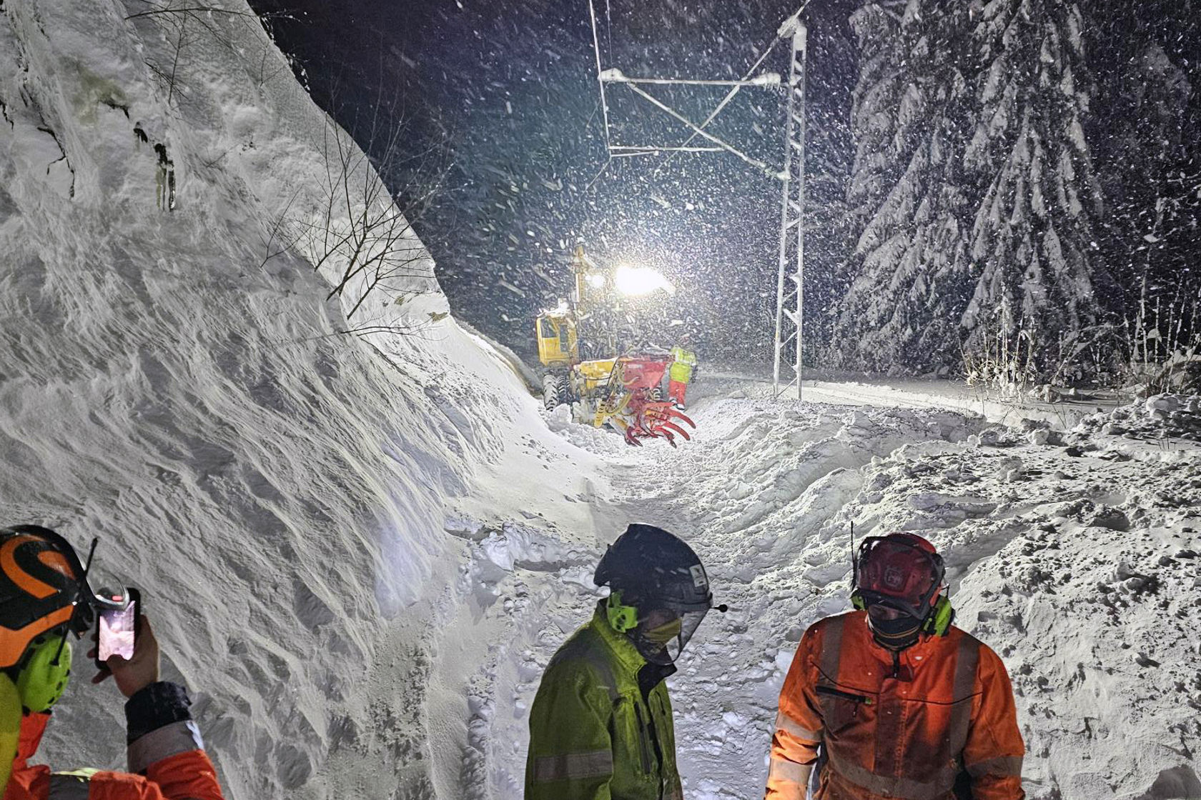 Folk i arbeidstøy står i forgrunnen, mens en arbeidsmaskin lyser opp himmelen der det snør kraftig.