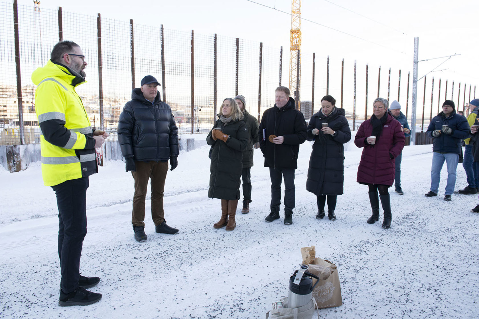 Gruppe mennesker med kaffe, gjerde og byggekran, vinter.