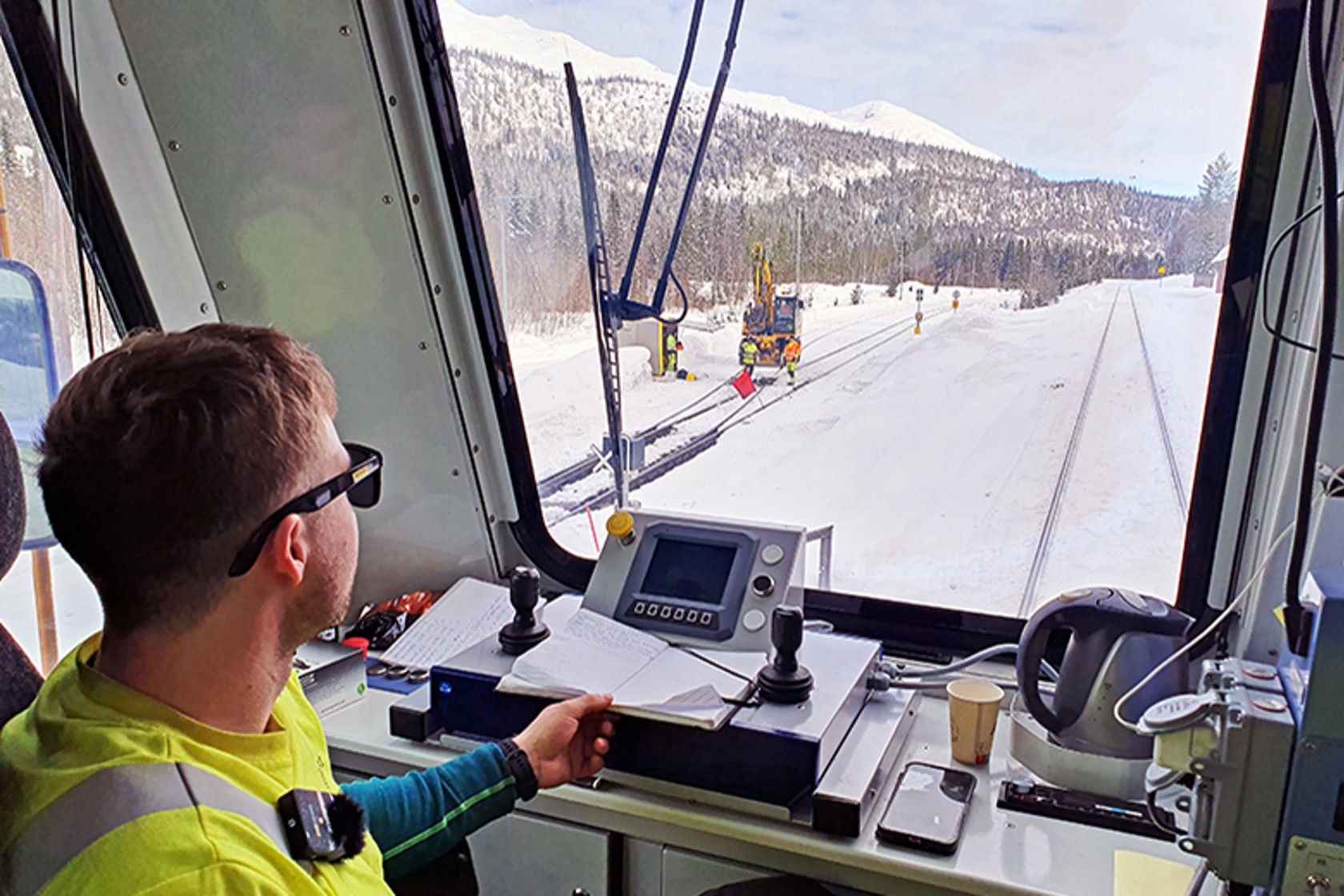 Mann sitter ved spakene i et arbeidstog med utsikt mot skog og snødekte fjell.